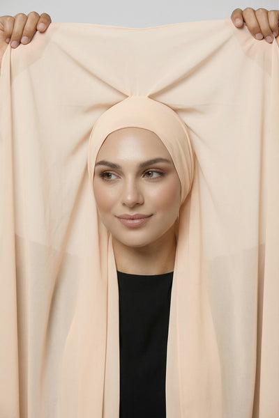 Model holding shell beige Nuramira chiffon hijab attached to undercap, showing full coverage and soft drape