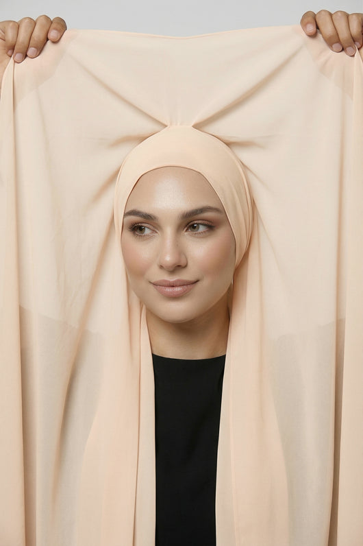 Model holding shell beige Nuramira chiffon hijab attached to undercap, showing full coverage and soft drape