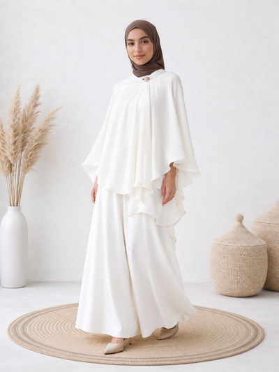 Front-facing woman wearing the Elara™ Pleated Cape Set, showcasing the flowing pleated cape, gold brooch detail, and wide-leg matching pants.