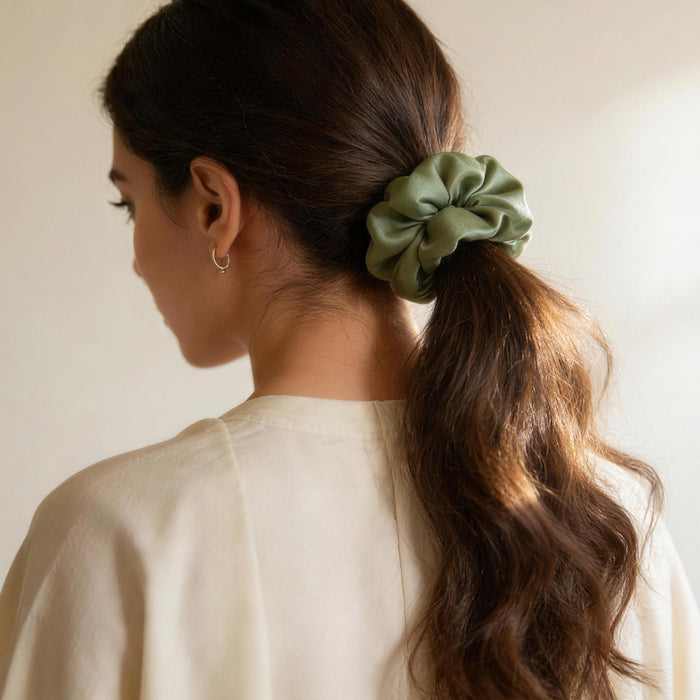 Rear view of a woman with long brunette hair wearing a lustrous cardamom green silk scrunchie holding a ponytail, styled with an off-white blouse.