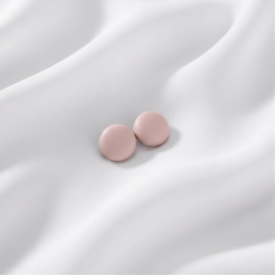 A pair of matte, soft dusty pink round magnetic hijab pins resting together on a draped white silk fabric.