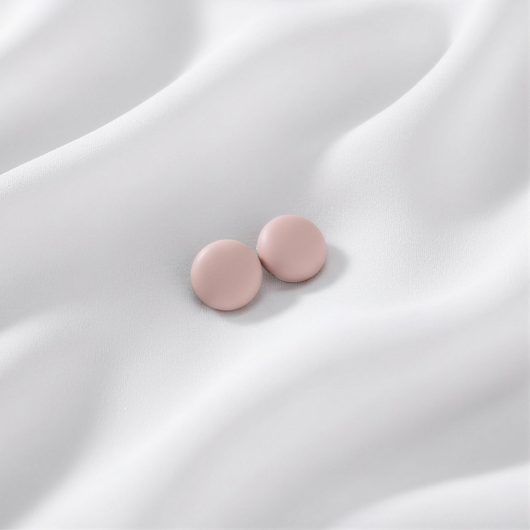 A pair of matte, soft dusty pink round magnetic hijab pins resting together on a draped white silk fabric.