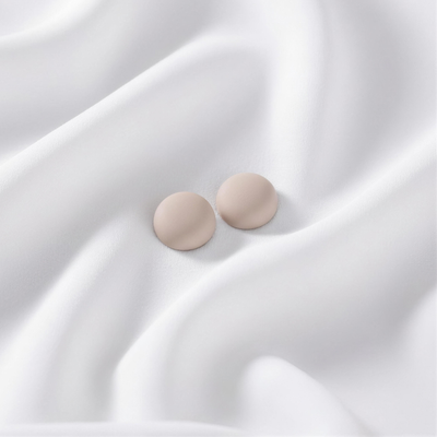 A pair of matte, neutral beige-sand round magnetic hijab pins placed on a smoothly draped white fabric.
