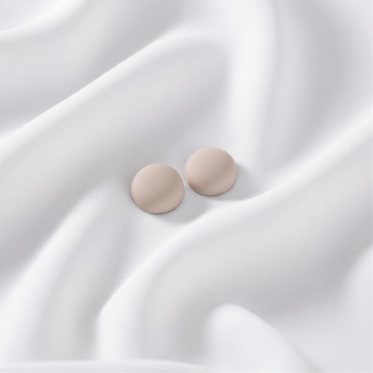 A pair of matte, neutral beige-sand round magnetic hijab pins placed on a smoothly draped white fabric.