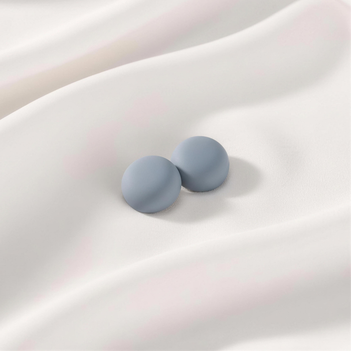 A pair of matte, slate blue-gray round magnetic hijab pins sitting side-by-side on a draped white silk fabric.