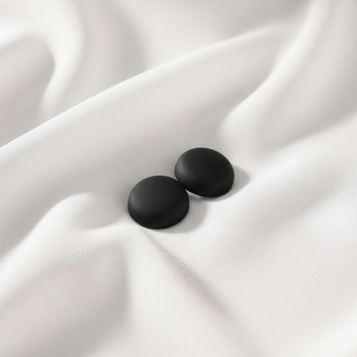 A pair of matte, sleek black round magnetic hijab pins resting together on a smooth, draped white fabric.