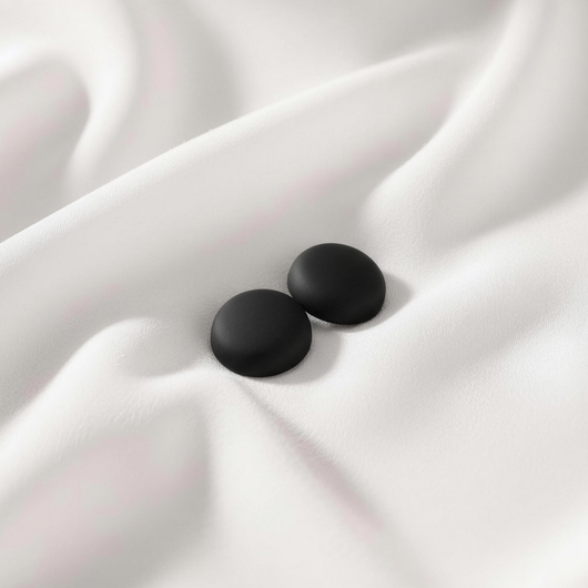 A pair of matte, sleek black round magnetic hijab pins resting together on a smooth, draped white fabric.