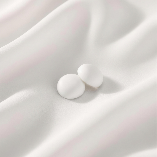 A pair of matte, clean white round magnetic hijab pins displayed on a softly folded white silk backdrop.