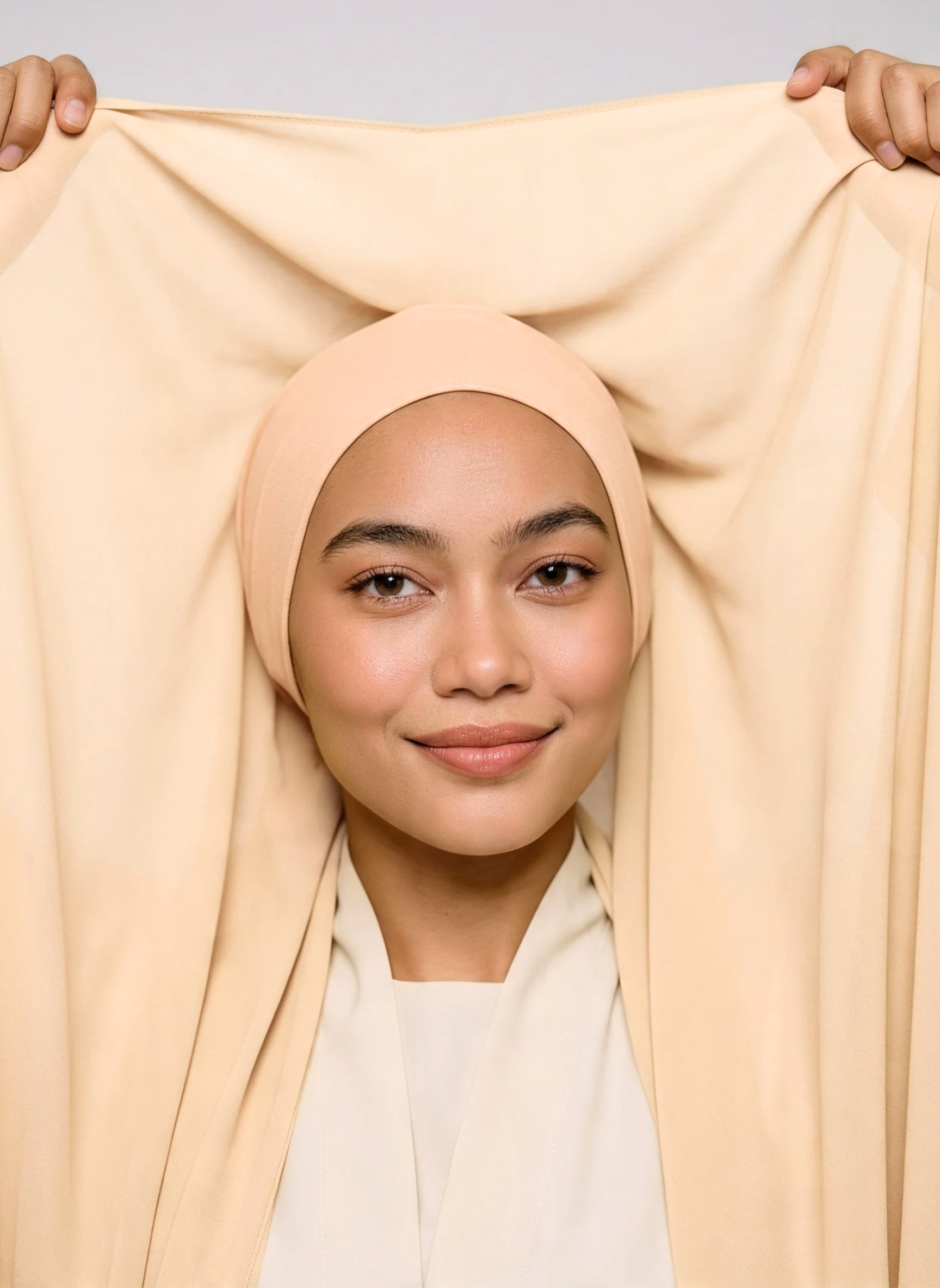 Model holding shell beige Nuramira chiffon hijab attached to undercap, showing the length of the chiffon and cotton breathable undercap