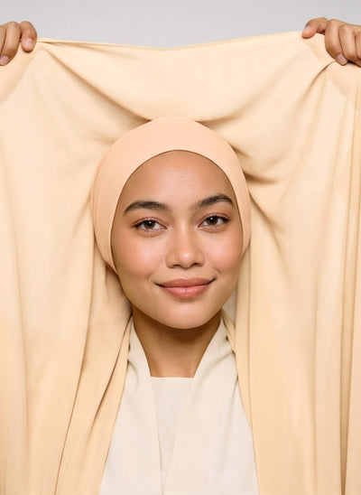 Model holding shell beige Nuramira chiffon hijab attached to undercap, showing the length of the chiffon and cotton breathable undercap