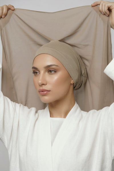 Woman lifting the new Nuramira instant chiffon hijab in praline beige attached to upgraded undercap with mesh panel, adjustable tie, and velvet grip in white studio